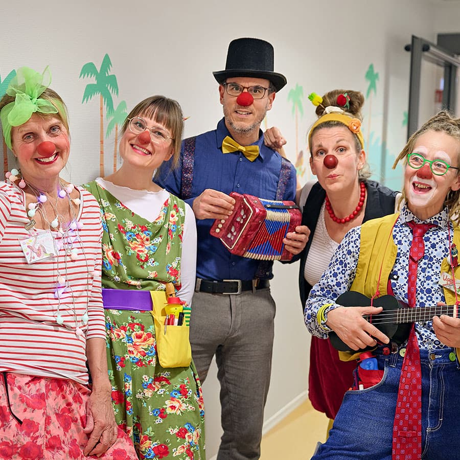 Eine Gruppe von kostümierten Klinikclowns steht im Flur der Kinderklinik und schaut lachend in die Kamera.
